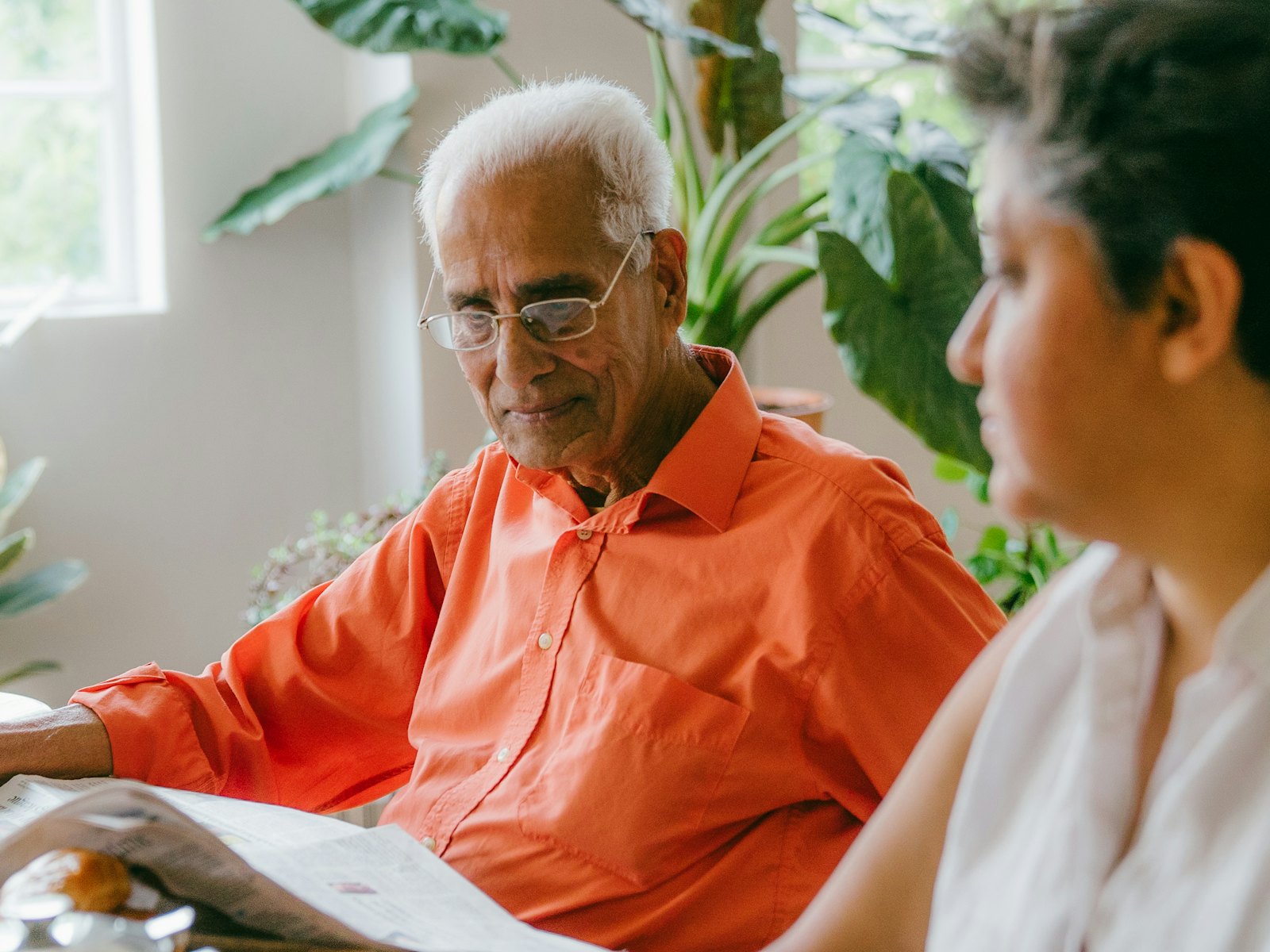 Un père âgé lit le journal à la table familiale, à côté de sa fille adulte, dans une pièce claire pleine de plantes